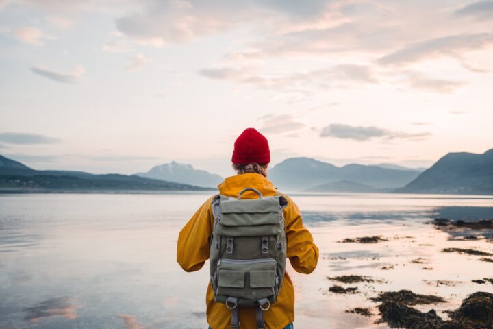 Back view of male tourist with rucksack standing on coast in front of great mountain massif while journey. Man traveler wearing yellow jacket with backpack explore scandinavia nature. Wanderlust outdoor lifestyle
